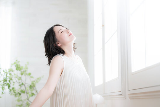 窓辺で朝の光を浴びながら深呼吸の伸びをする女性の健康ライフスタイルイメージ