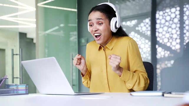 Excited young businesswoman in big wireless headphones watching sports match on laptop at office desk. Happy female employee cheering for auction bids and celebrating success showing achievement.