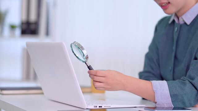 Asian businesswoman holding magnifying glass examining laptop screen searching information research analysis data verification inspection quality control
