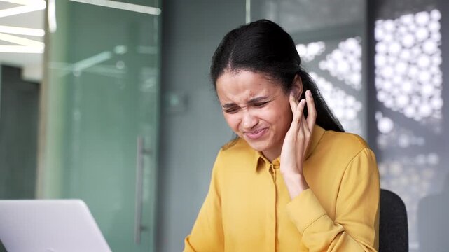 Close up of female hand touching and massaging sore ear at office desk. Tired businesswoman suffering from otitis media, earache and sharp discomfort while sitting at modern business workplace.