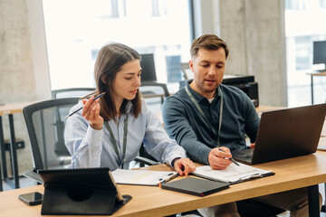 Fototapeta premium Two professionals engaged in discussion at a modern office workspace, one woman holding a pen and the man pointing at a laptop screen with documents on the table