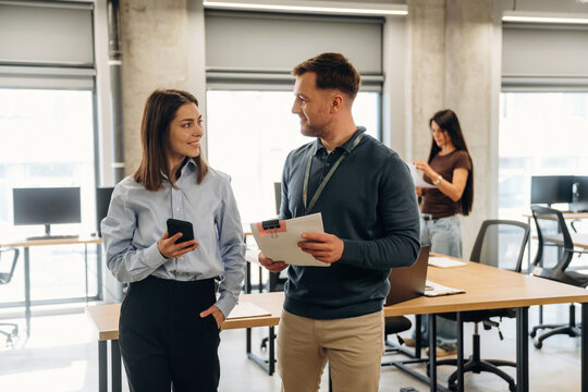 Young Caucasian woman and man engaged in conversation inside modern office space with desks and computers, another woman using smartphone in background