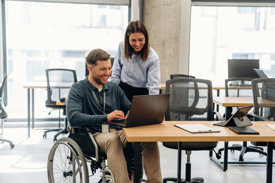 Male office worker in wheelchair using laptop while female colleague assists him in modern workspace with large windows and wooden desks