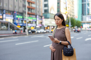 Fototapeta premium Pregnant asian woman checking her mobile phone on city street