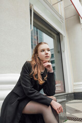 Fototapeta premium A beautiful young stylish and fashionable girl in a black coat walks through the city streets