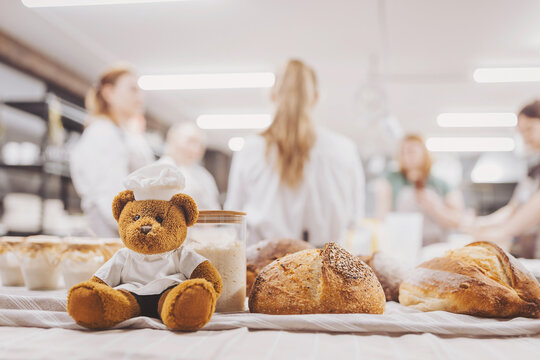 Baker prepares fresh sourdough bread while a teddy bear chef observes in a bustling bakery kitchen with people in the background