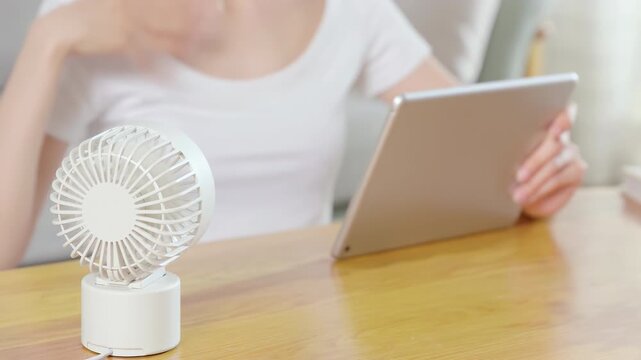 Unwell young woman employee sitting and working with laptop tablet using ventilator portable handheld mini fan blowing, stressed annoyed girl at home breathing fresh cool air, summer heat weather