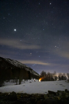View of people gathered around a glowing campfire in a snowy field under a clear starry night sky with the Orion constellation and dark mountains in Lakselvbukt, Troms, Norway.