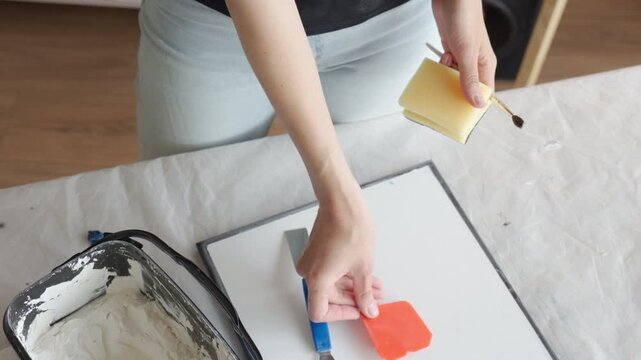Woman arranges sponge brush and palette knife on canvas at a protected work table. Concept of art supplies DIY painting setup and handmade decor.