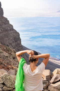 Woman standing on cliff pulling hair back, looking at blue ocean waves and volcanic landscape, embracing travel and freedom