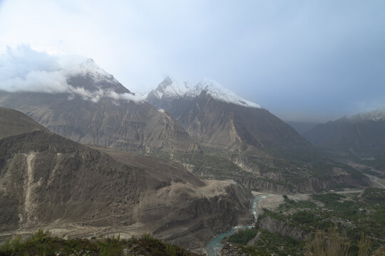 View of snow-capped mountain peaks towering over a winding river valley with rocky slopes and scattered greenery under a cloudy sky in Gilgit-Baltistan, Pakistan.