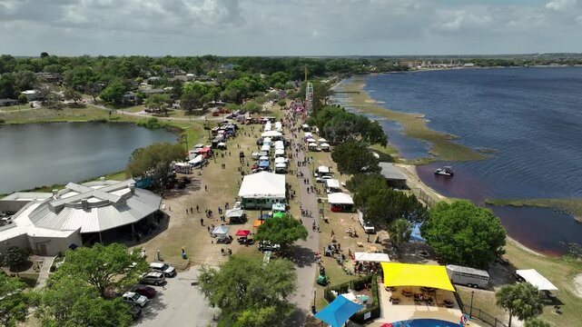 Cinematic bird's-eye shot of Clermont's Pig on the Pond, capturing carnival rides, vendor booths, and festival-goers gathered along the scenic shores of Lake Minneola.