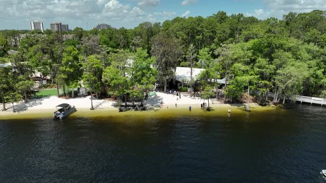 Aerial drone view of Paradise Cove on Lake Bryan in Orlando, Florida, featuring a sandy lakeside beach, docked pontoon boat, and lush tropical greenery framing the shoreline.