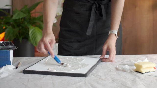 Woman makes textured picture at home using canvas plaster paste and tools. Concept of DIY home decor handmade picture and creative hobby.