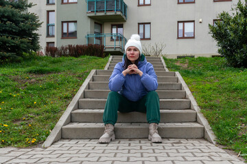 Woman in winter hat and blue puffer jacket sits on concrete stairs near an apartment building, resting with hands together in a quiet urban outdoor setting. © Volha