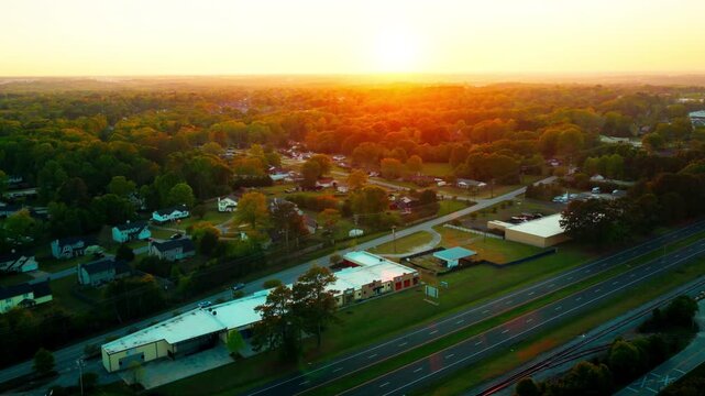 Golden hour drone aerial over Greenville, South Carolina suburbs with sun centered on horizon, backlit residential streets, divided highway, parallel railway, and vast forest canopy stretching beyond.