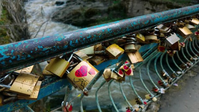 Close-up view of numerous "love locks" attached to a rustic bridge railing in Borjomi, Georgia. Many locks feature heart designs, symbolizing affection and commitment, with the blurred river backgr...