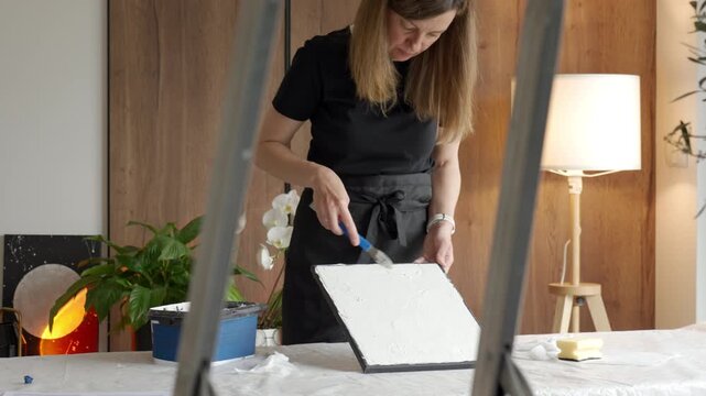 Woman makes textured picture at home spreading plaster paste on canvas with a palette knife. Concept of DIY wall decor handmade art and creative hobby.