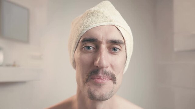 Funny mustached man in bathroom with towel on head smiles and squints during grooming routine