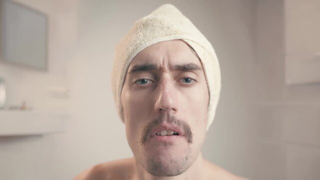 Funny mustachioed man in bathroom with towel on head looking questioningly and frowning