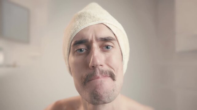 Uncertain mustached man in bathroom with towel nods hesitantly, gestures doubt, contemplates with varied facial expressions and cheek puffing