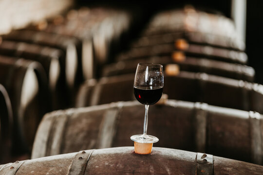 View of a glass of red wine resting on a wooden barrel in a traditional wine cellar with rows of aging oak casks Coka, Vojvodina, Serbia.