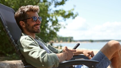 Obraz premium A man is sitting in a chair by the water, smiling and holding a cell phone