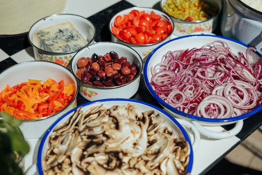 Bowls of sliced onions, mushrooms, peppers, tomatoes, olives, cheese, and dough arranged on a checkered table for pizza preparation