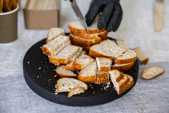 Gloved hand slicing fresh bread into sample pieces on a black wooden serving board with crumbs scattered on a tablecloth