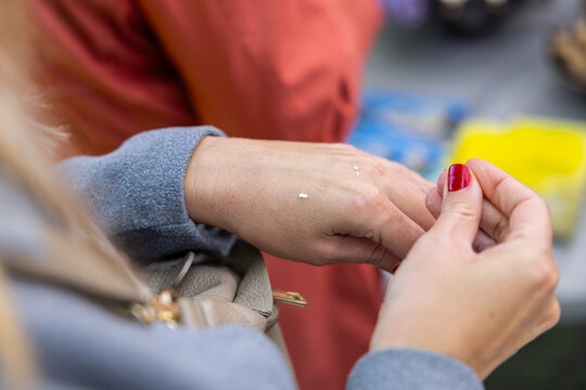 Close-up of hands applying small dots of cream on a wrist during product sampling, with red manicure and blurred market background