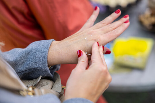 Close-up of hands applying small dots of cream on a wrist during product sampling, with red manicure and blurred market background