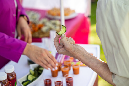 At an outdoor market, a person holds a cucumber slice near tomato juice samples, cut cucumbers, and tasting cups on a bright table