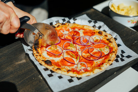 Hand using a pizza cutter to slice a wood-fired pepperoni pizza topped with red onion and peppers on branded parchment paper