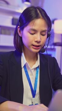 Smiling young Asian customer service representative wearing microphone headset and working on laptop in modern office.