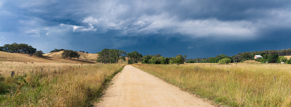 Low angled panorama view of a gravel road through long grass with dark storm clouds overhead