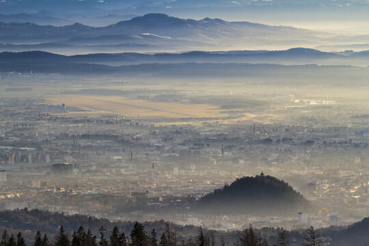 View of the historic city of Graz with the Schlossberg hill and clock tower emerging from morning mist against a backdrop of distant mountains and the airport Graz Thalerhof, Styria, Austria.