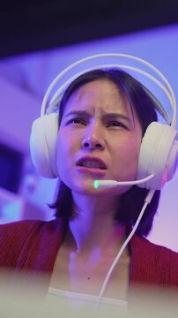Excited young Asian girl playing online computer game with headset and microphone at home, esports and live streaming concept.