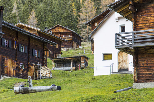View of traditional wooden alpine houses and a white chapel nestled on a green hillside with a dense forest background in Innervillgraten, Tyrol, Austria.