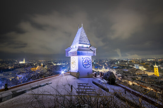 View of the illuminated Uhrturm clock tower covered in snow overlooking the city lights at night in Graz, Steiermark, Austria.