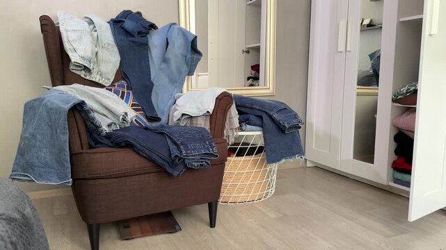 Folded jeans and clothes on armchair beside laundry basket with open wardrobe door during closet decluttering camera pan right to left