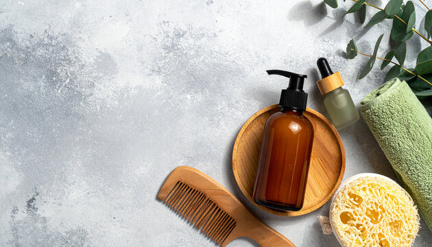 Spa Essentials: Natural beauty products, including a wooden comb, luffa sponge, amber glass bottle with pump, essential oil dropper, and green towel, are artfully arranged on a textured gray surface.