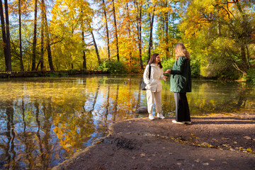 Fototapeta premium Two woman talking in autumn park near pond. Friends walking outdoors on bright fall day. Conversation in nature, leisure time lifestyle, seasonal recreational activity, community.