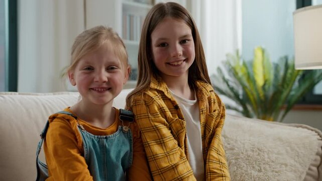 Portrait two happy Caucasian sisters siblings smiling joyful relaxing on couch looking at camera at home cheerful children little girls daughters kiss smile posing in living room together good family