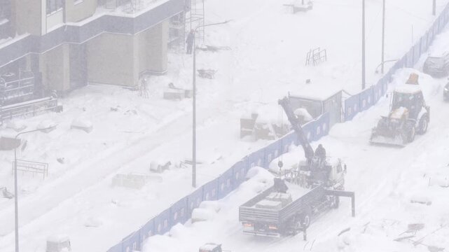 Truck-mounted crane operating at construction site during heavy snowfall in harsh winter conditions, loading and unloading building materials