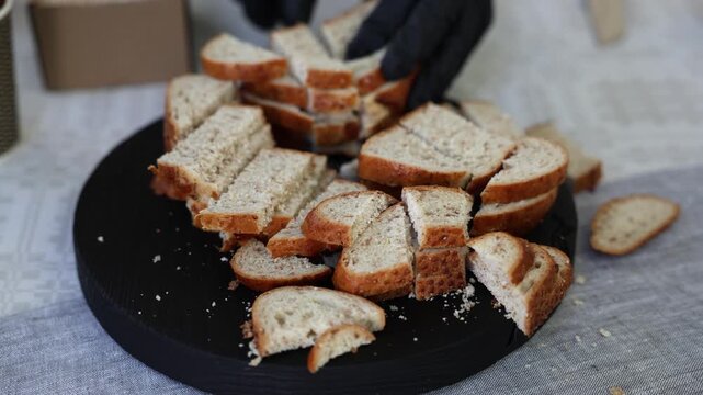 Close-up of sliced bread pieces on a black serving tray, with a gloved hand cutting fresh loaf on a patterned tablecloth