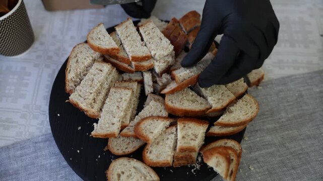 Close-up of sliced bread pieces on a black serving tray, with a gloved hand cutting fresh loaf on a patterned tablecloth