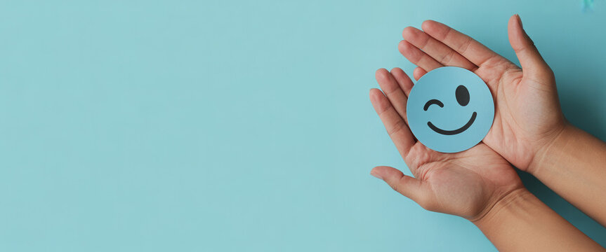 A photograph of two hands gently holding a light blue circular smiley face against a pale blue background palms fingers object happy