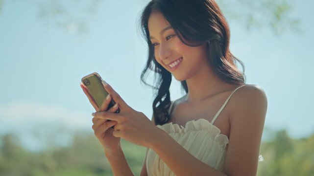 Happy young Asian woman in white dress smiling while using smartphone outdoors in summer park, portrait of beautiful female typing online message, social media connection, lifestyle and technology.