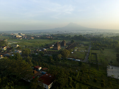 Aerial view of Plaosan Lor Temple complex surrounded by lush green fields and residential houses with Mount Merapi in the background Kecamatan Prambanan, Jawa Tengah, Indonesia.