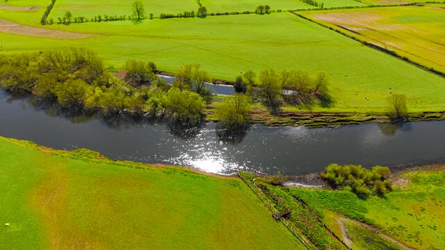 Sunlit river segment cutting through green farmland with small drainage channel visible. Aerial drone view water reflections grassy banks trees and agricultural plots forming landscape. Irrigation